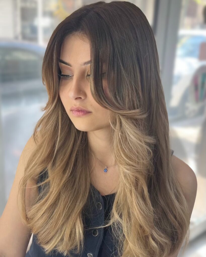 A woman with long dark hair transitioning into caramel blonde ombre with sweeping face framing layers and curtain bangs.