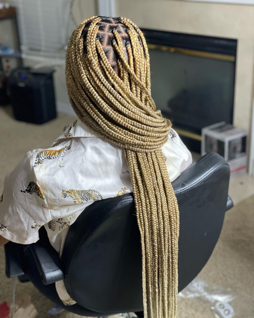 A back view of a person with long thick knotless braids in a bright blonde color styled in a low ponytail wrap.