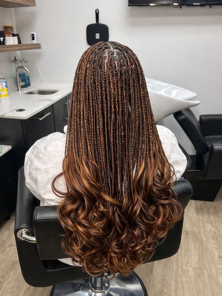 A woman sits in a salon chair displaying thin brown and copper knotless braids that end in large bouncy ringlet curls.