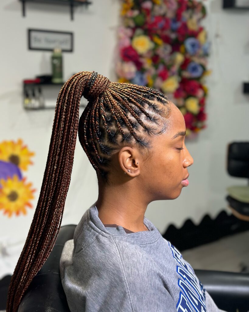 A woman with side profile showing honey blonde and brown knotless braids styled into a high sleek ponytail.
