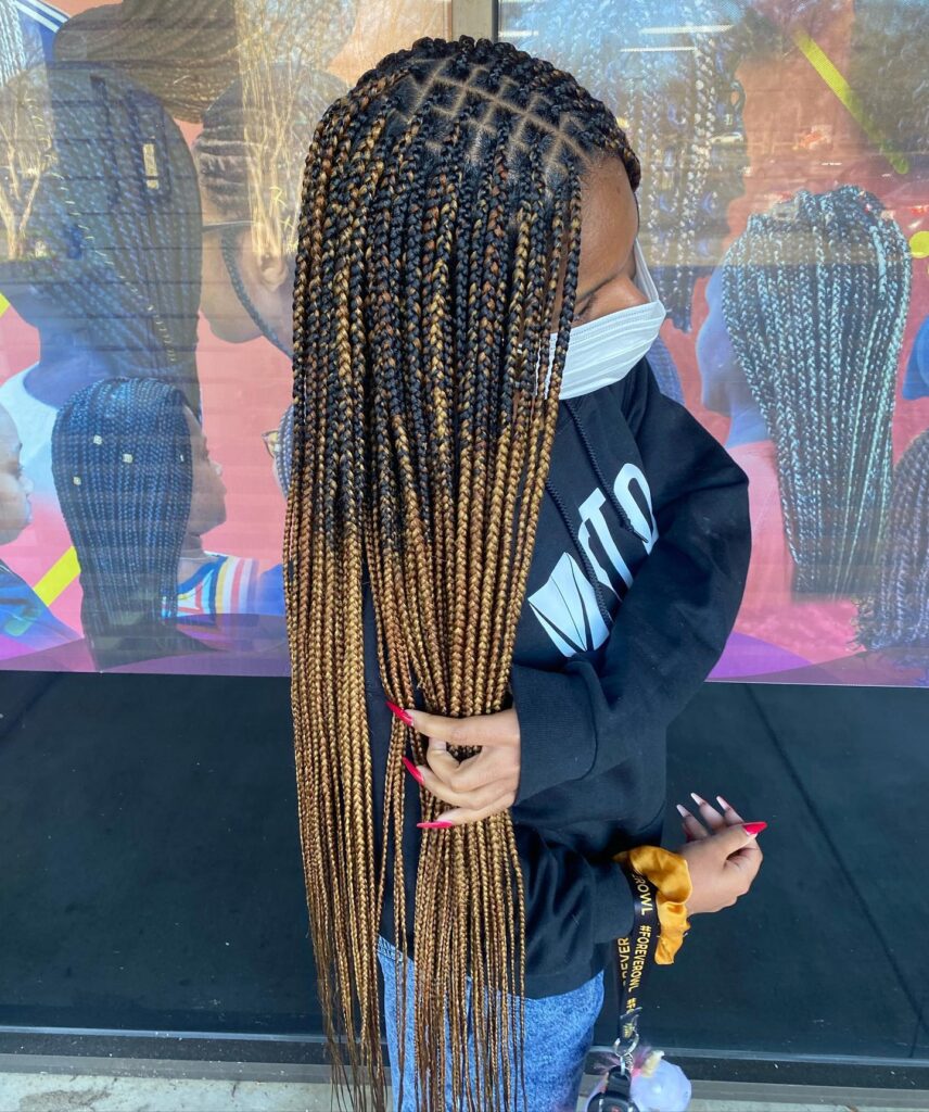 A woman stands sideways showing off her extra long knotless braids that fade from black to a bright caramel blonde.