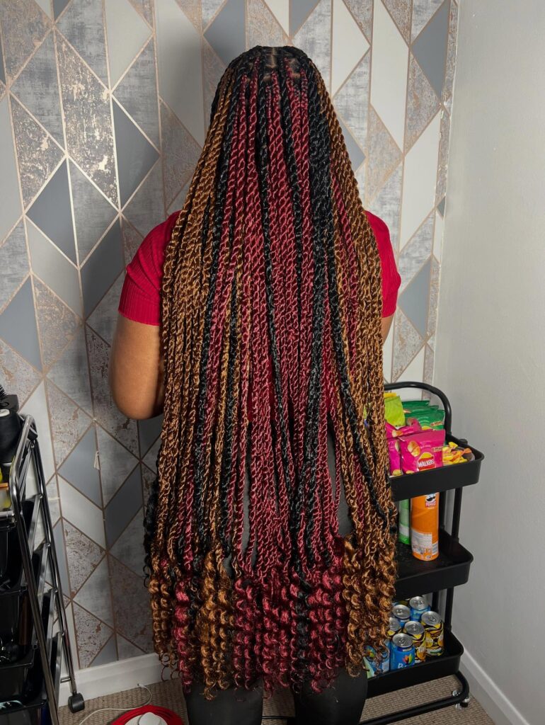 A back view of a woman with long knotless braids in a mix of copper red and black colors ending in thick voluminous curls.