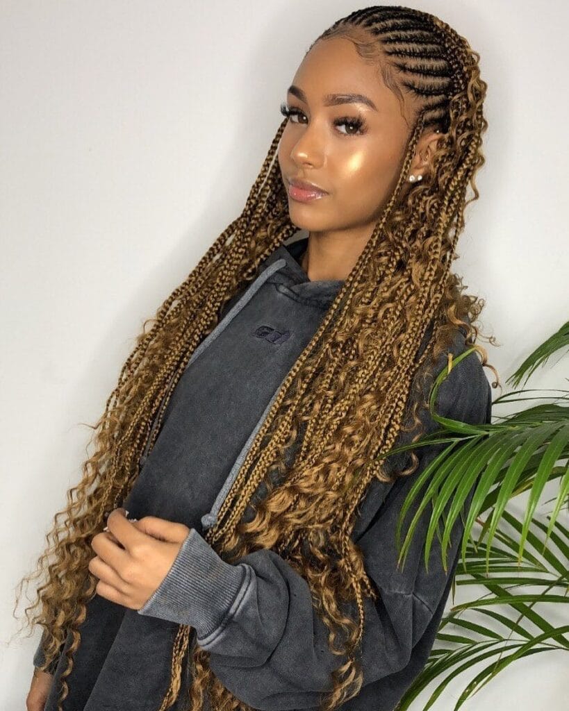 A young woman posing with long honey blonde braids that feature intricate cornrows on top and loose wavy curls throughout the ends.