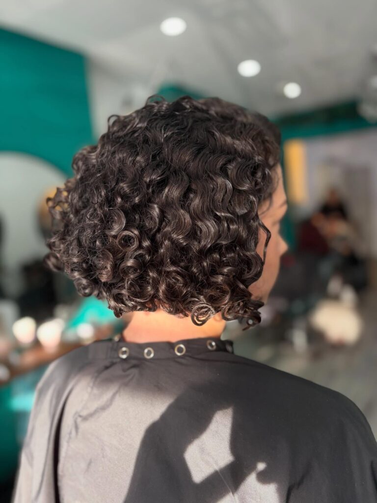 A person from the back with a very shiny, neck-length curly bob in a dark brunette color, showing defined ringlets.