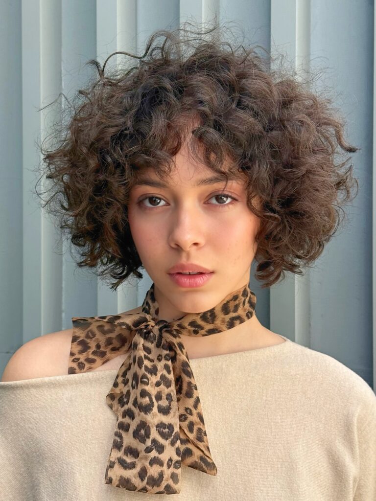 A young woman with a voluminous chin-length ash brown curly bob and soft wispy bangs wearing a leopard-print neck scarf.