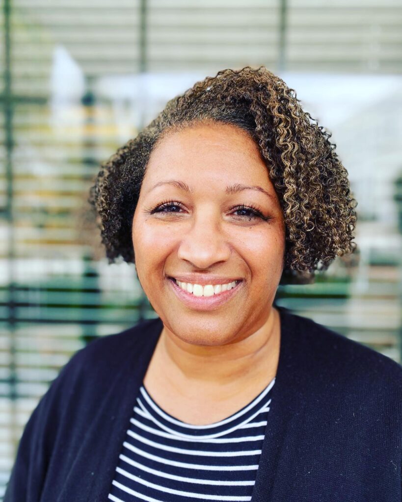 A smiling woman with a chin-length bob featuring tight natural coils and sandy blonde highlights.