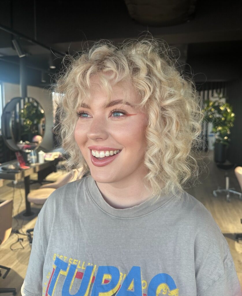 A smiling woman with a voluminous neck-length champagne blonde curly bob and curly bangs.