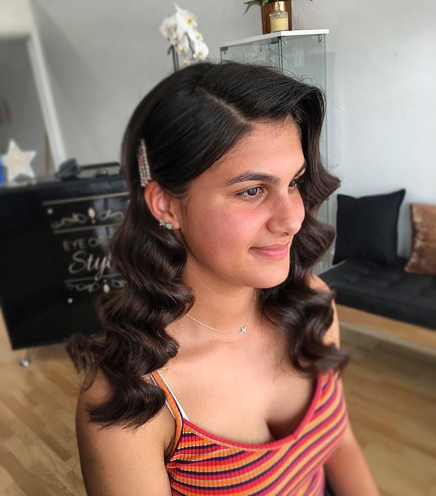 A young woman with deep brunette hair styled in glossy vintage waves with a side part and a sparkly hair clip.