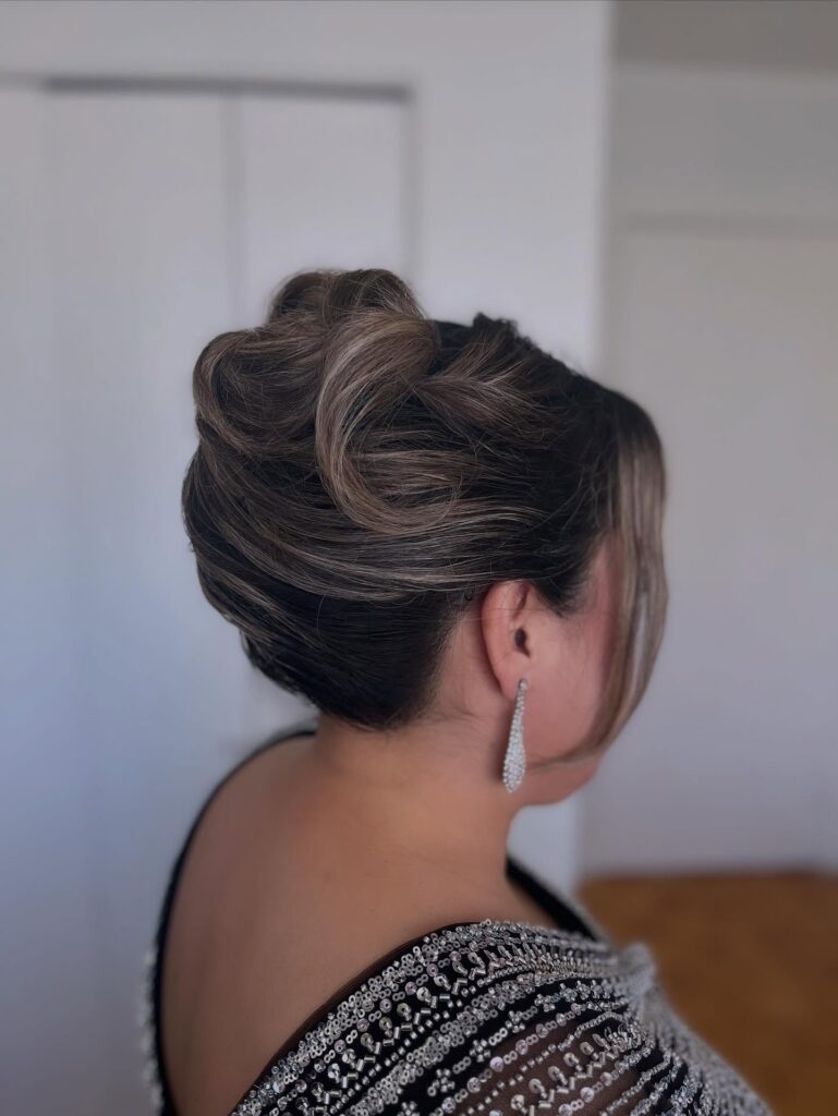 A woman viewed from the back, showing an intricately sculpted updo with ash blonde highlights and soft textured rolls.