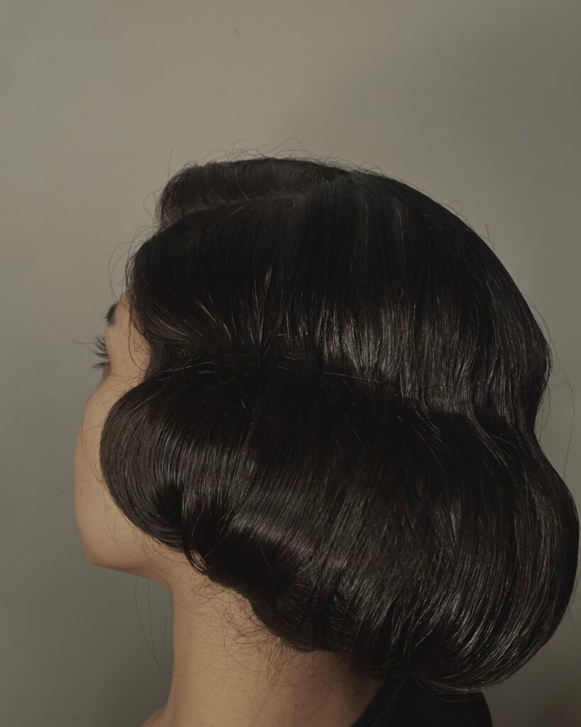 A side view of a woman with dark black hair styled into a voluminous faux bob with glossy retro finger waves.