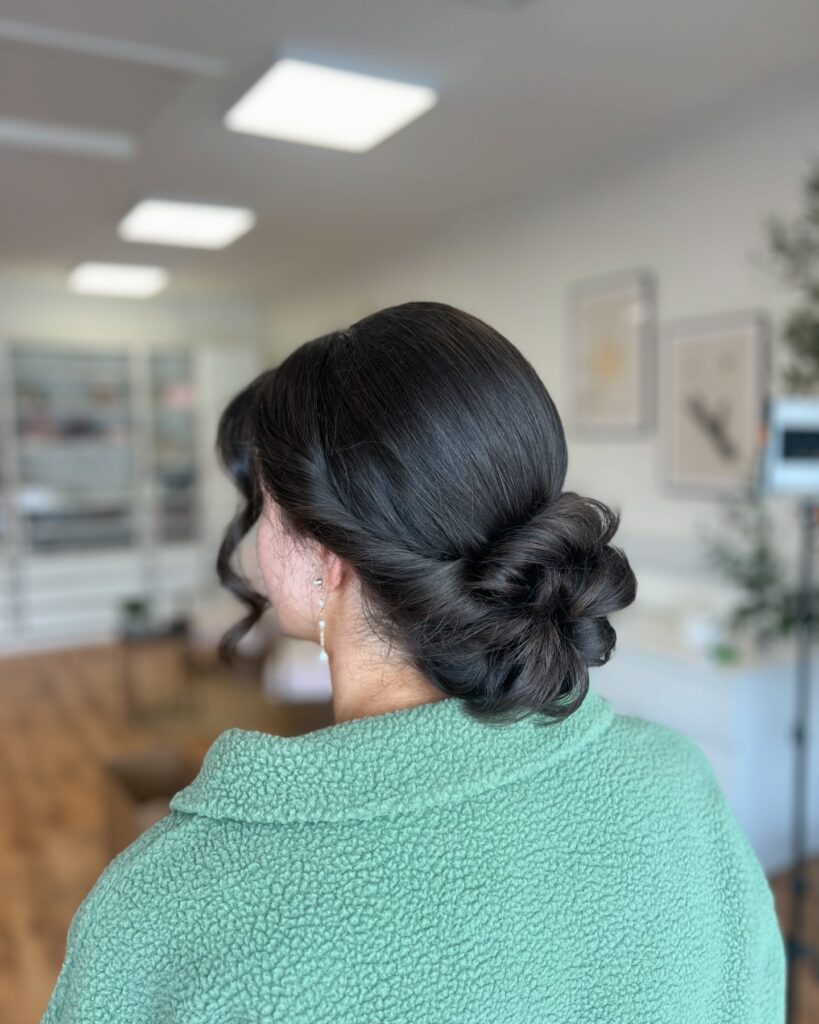 A woman viewed from the back, showing a textured low bun in dark hair with soft curled tendrils framing her face.