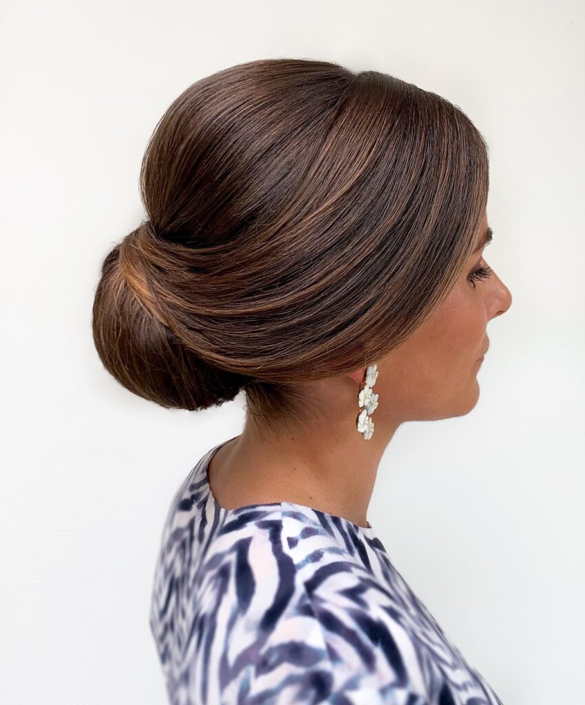 A side profile of a woman with chestnut brown hair styled in a voluminous, smooth low chignon with elegant swept-back layers.