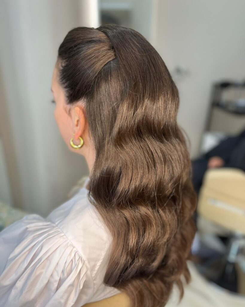 A woman viewed from the side wearing long brunette hair styled in glossy Hollywood waves with the front sections tucked back.