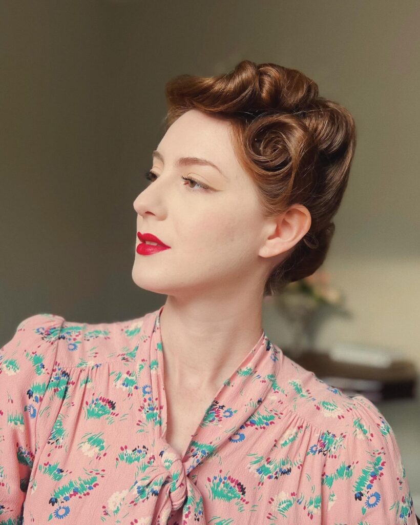 A woman with auburn hair styled in classic high-volume victory rolls and a polished vintage updo.
