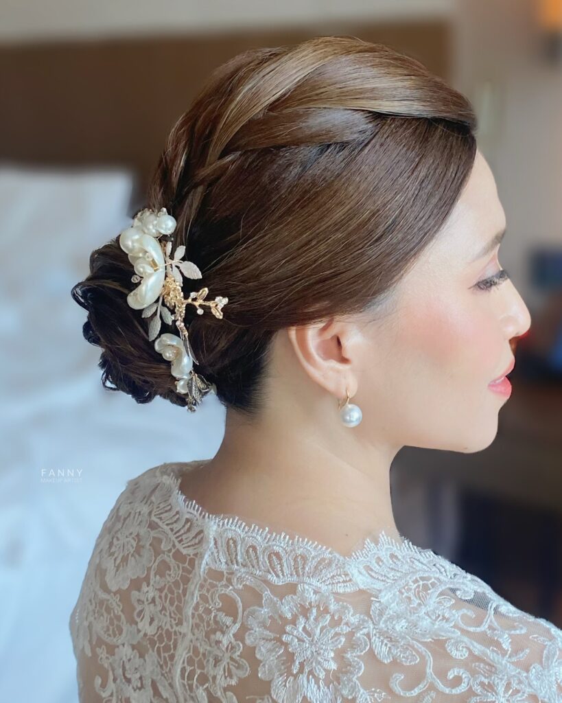 A chestnut brunette updo featuring a side braid and a low bun decorated with a large pearl and gold floral hair clip.