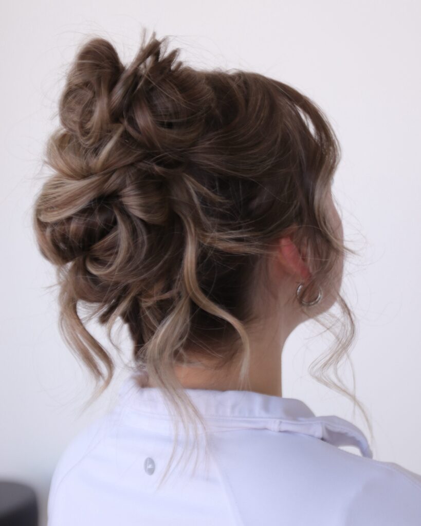 A messy high updo in ash-brown hair with blonde highlights and loose, wavy face-framing tendrils.