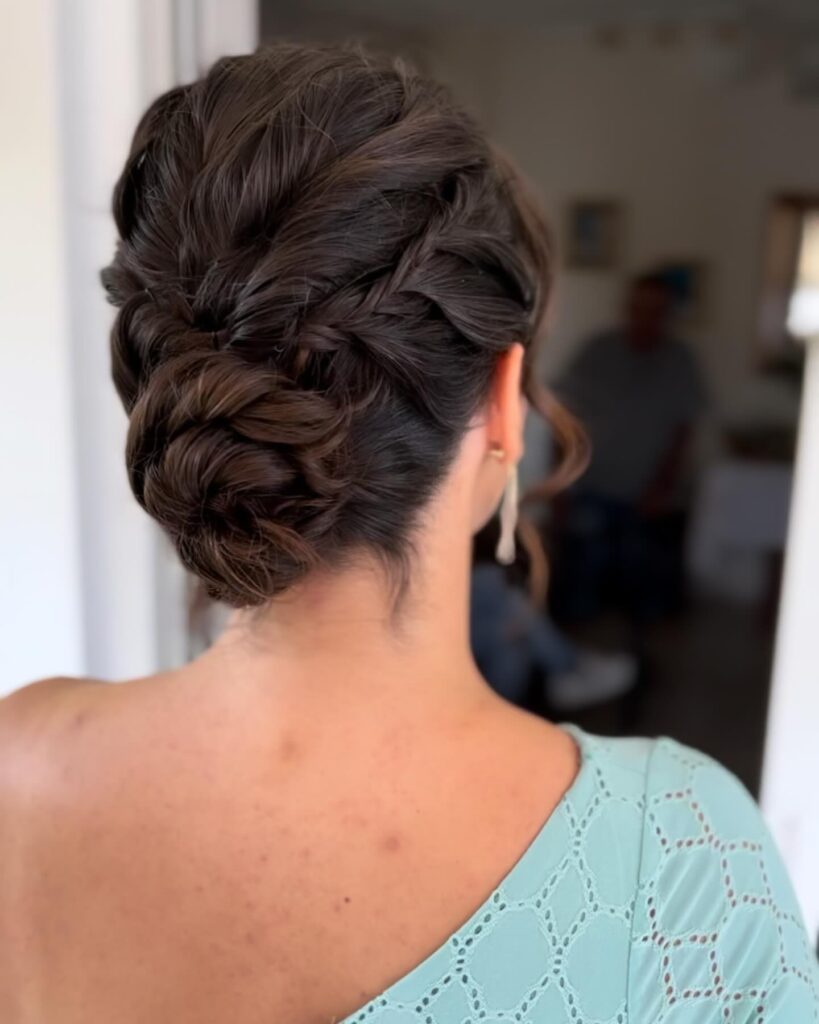 A textured low updo in dark brunette hair featuring an intricate side braid tucked into a soft chignon.