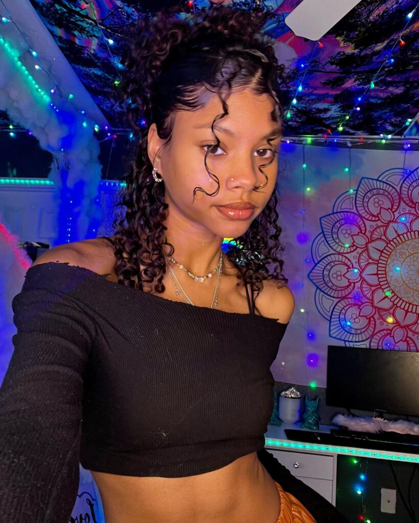 A young woman with dark curly hair in a half-up ponytail style with defined edges and two loose ringlets framing her face.