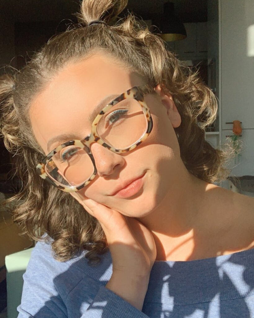 A young woman with light brown wavy hair styled in small topknot buns and wearing patterned glasses.