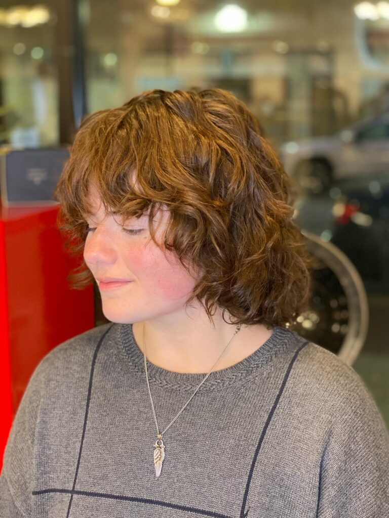 A person with shoulder-length wavy brown hair in a layered shag cut with wispy bangs.