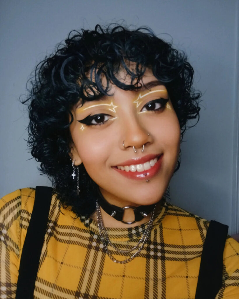 A smiling person with short, glossy, jet-black curly hair and edgy makeup.