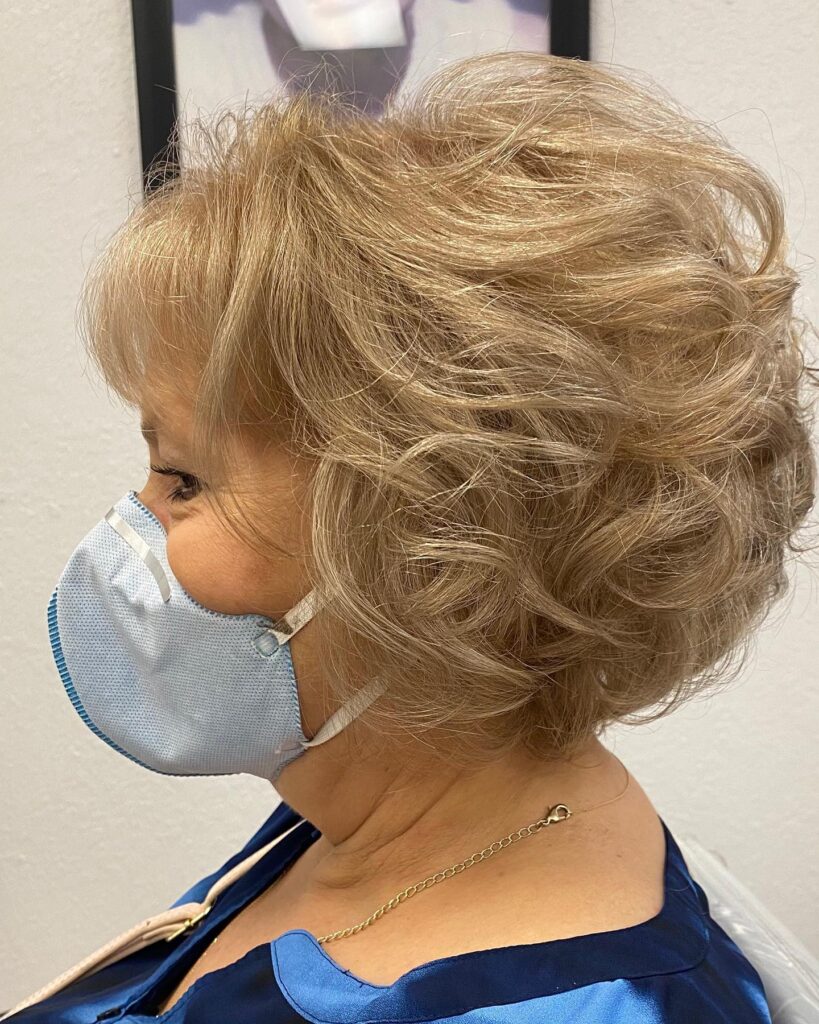 A woman viewed from the profile is sporting a chin-length voluminous champagne blonde bob with soft curls and wispy bangs.