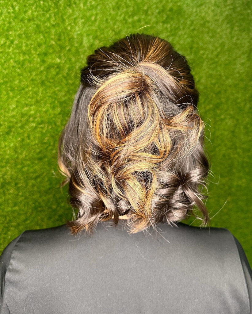 A back view of a woman with a dark brunette half-up hairstyle featuring bright golden blonde highlights and bouncy curls.