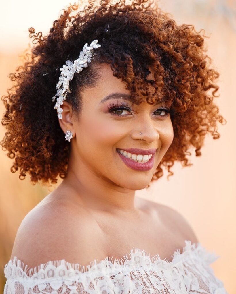 A smiling woman wearing a white lace dress with her natural coiled curls styled with a silver floral hair vine.