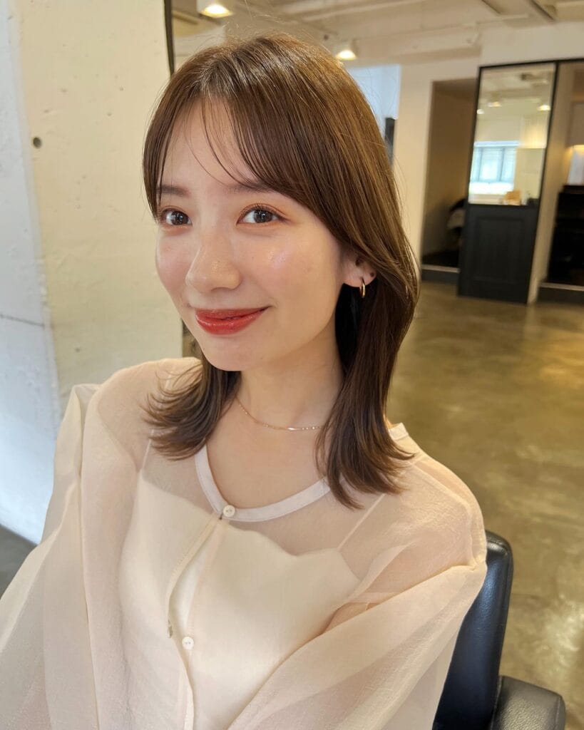 A young woman with a medium-length soft brown hairstyle featuring wispy bangs and ends that flip outward.