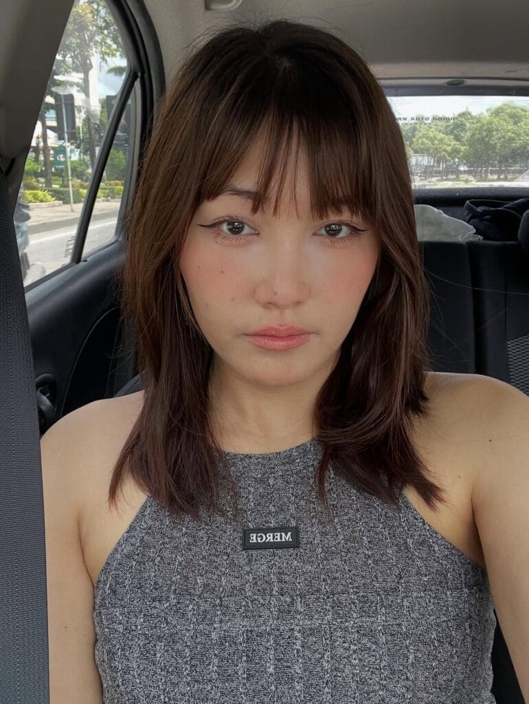 A woman with a medium-length chocolate brown shaggy haircut and thin wispy bangs is sitting in a car.