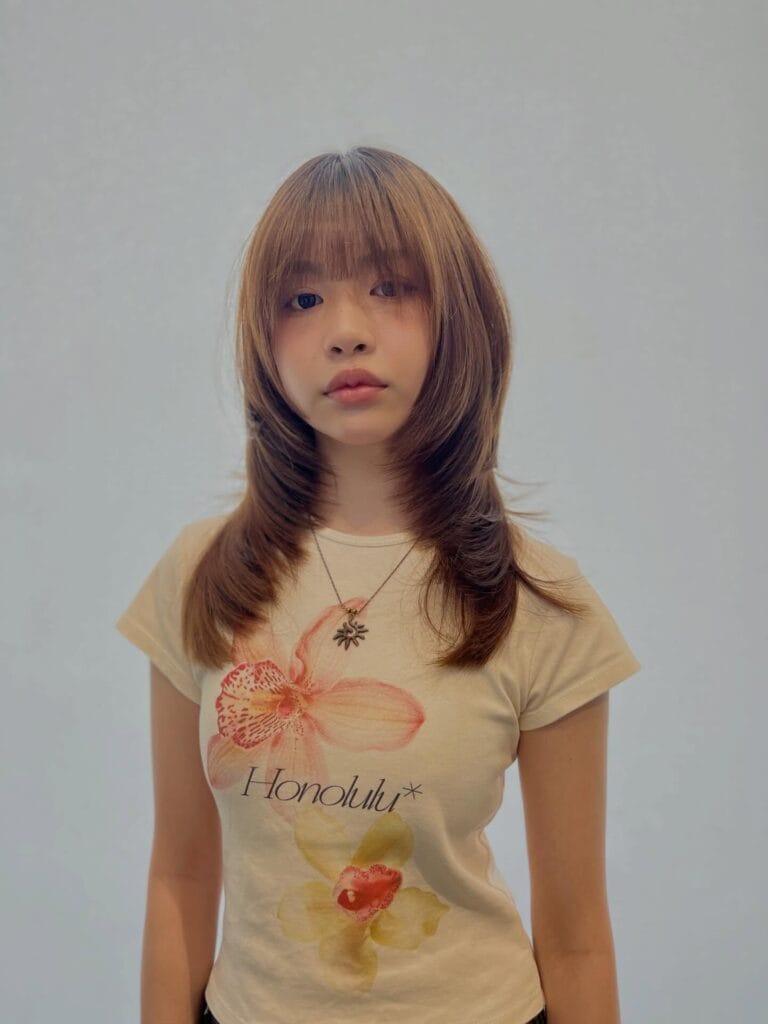 A young woman with a medium-length caramel brown butterfly haircut featuring thick bangs and tiered layers.