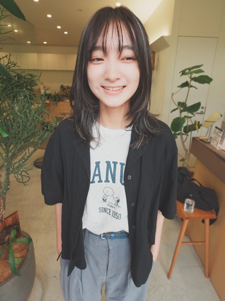 A smiling woman with a layered, dark, medium-length hairstyle and thin, fringed bangs, wearing a graphic t-shirt and an open black button-down shirt.