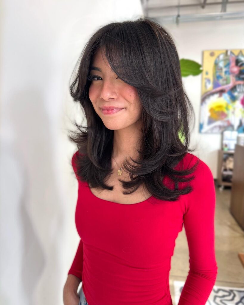 A smiling woman wearing a red top with a voluminous dark medium-length hairstyle featuring butterfly layers and sweeping curtain bangs flipped outward at the ends.
