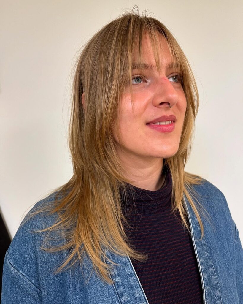 A woman with a medium-length golden blonde shag hairstyle featuring thin see-through bangs and textured layers.