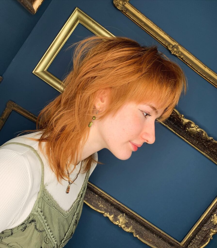 A woman in profile with a bright copper medium-length wolf cut featuring messy layers and thick straight bangs against a blue wall with gold frames.