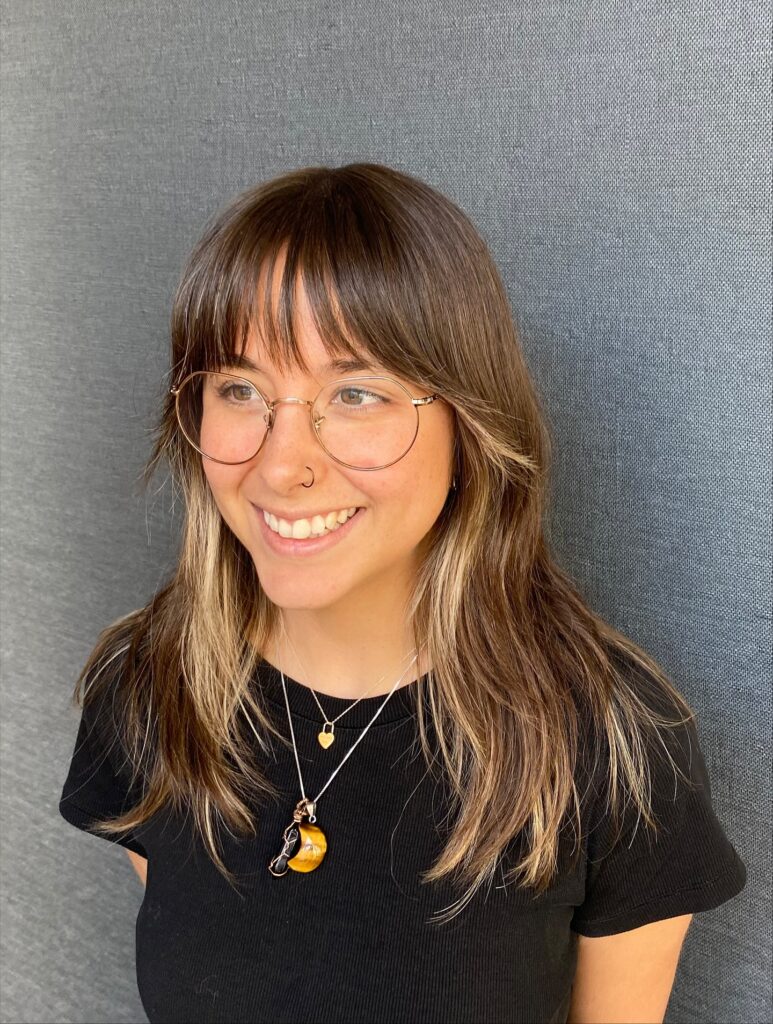 A smiling woman with glasses wearing a medium-length brunette hairstyle with blonde balayage highlights and a straight-across fringe.