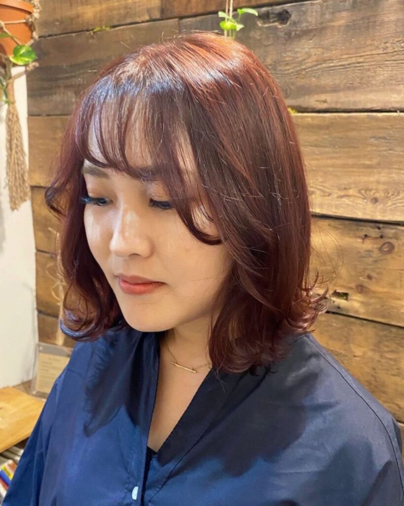 A woman with a medium-length auburn hairstyle featuring bouncy curled-in ends and soft wispy bangs.