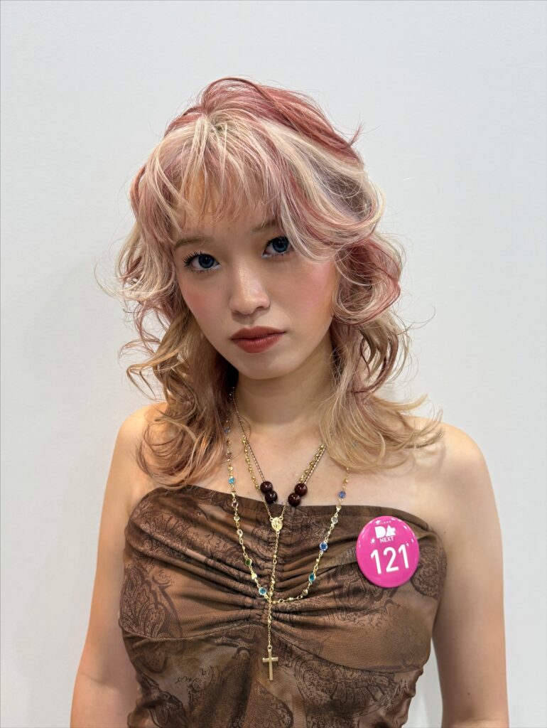 A woman with a medium-length shaggy hairstyle featuring a mix of pastel pink and blonde hair colors with curly layers and a wispy curled fringe.