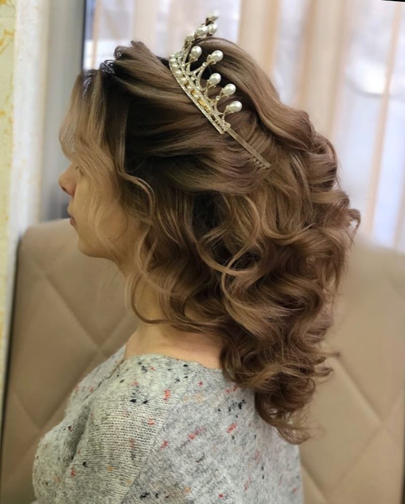 A side profile of a woman with medium-length curly brown hair styled in a side-swept look and topped with a pearl tiara.