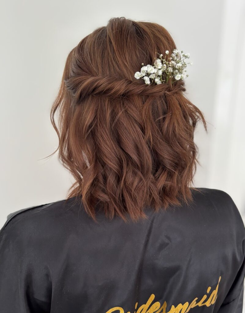 A back view of a woman with medium auburn hair styled in messy waves with a rope twist decorated with white baby's breath flowers.