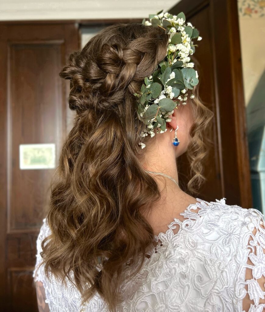 A woman with medium-length wavy brown hair styled in a complex braided half-up look, adorned with fresh eucalyptus and small white flowers.