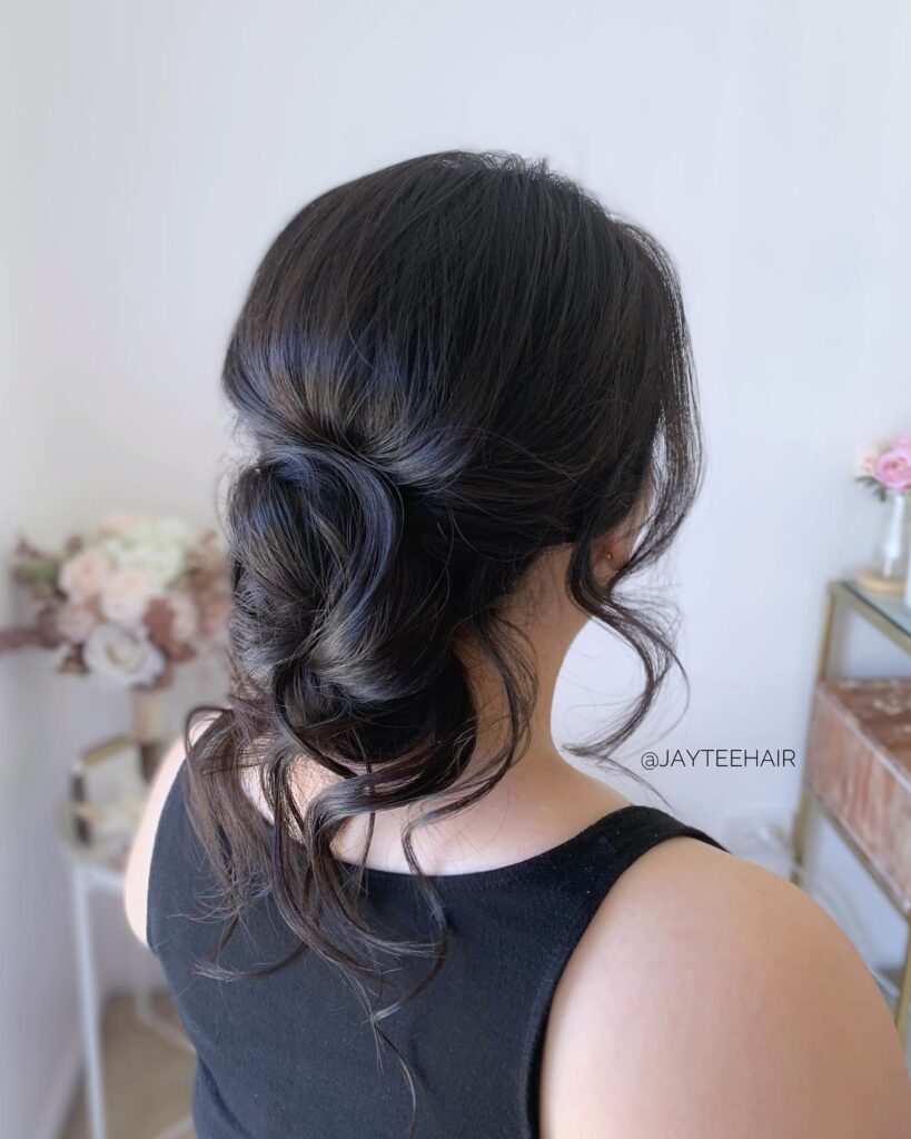 A back view of a woman with dark hair styled in a textured low bun with soft, loose curls framing her neck.