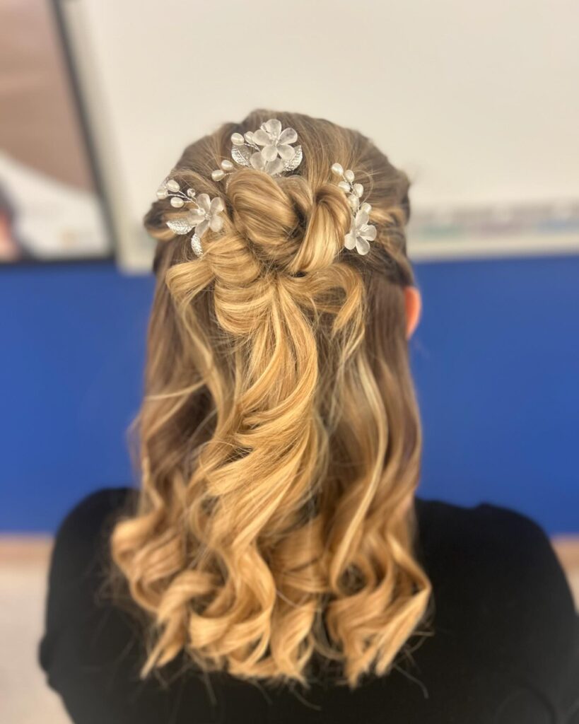 A rear view of a woman with medium-length blonde hair styled in a knotted half-up look with defined curls and floral pearl hair accessories.