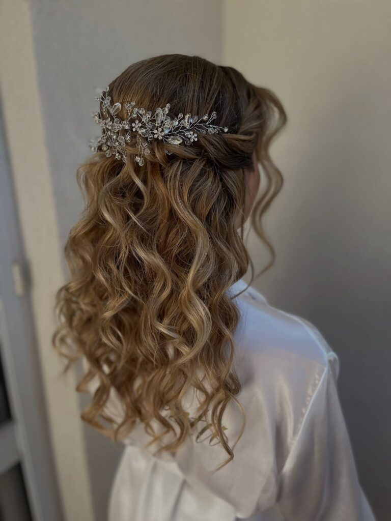 A back view of a woman with medium-length blonde curly hair styled in a half-up look with a large crystal hair vine accessory.
