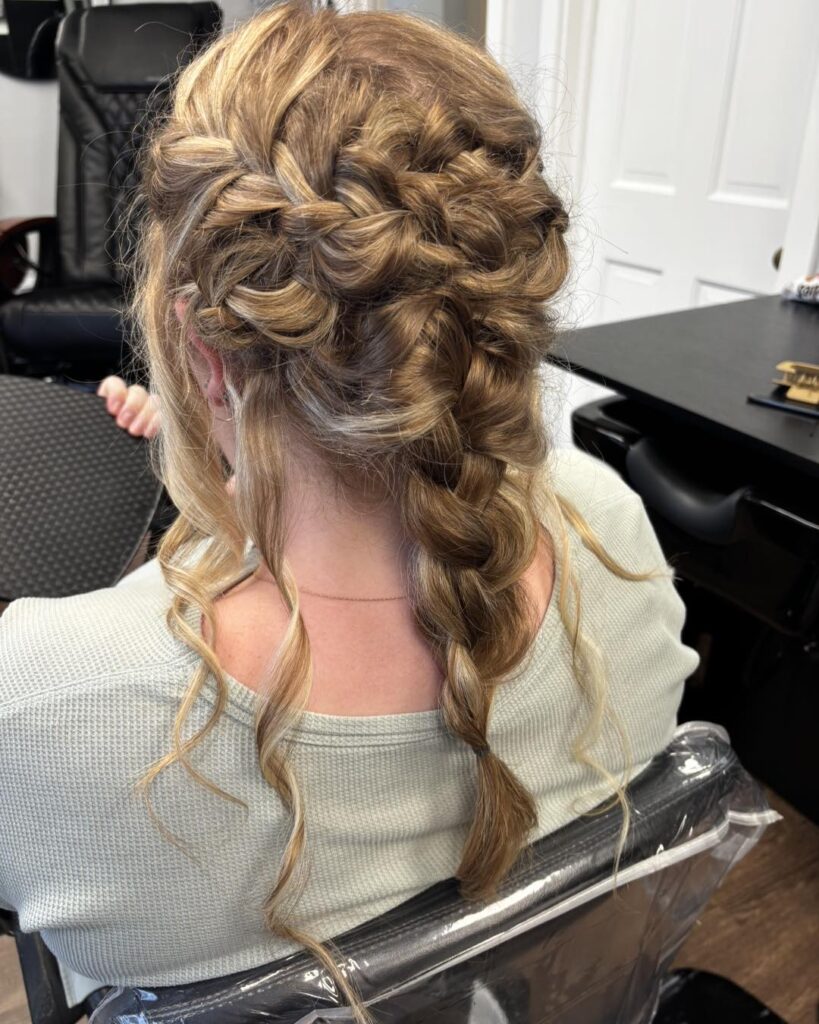 A back view of a woman with medium-length blonde hair styled in a thick loose braid with several soft curled tendrils framing the neck.