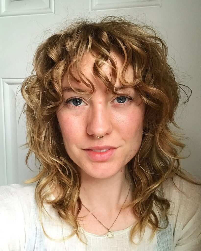 A woman with a medium length wavy shag haircut with curly fringe and soft layers.