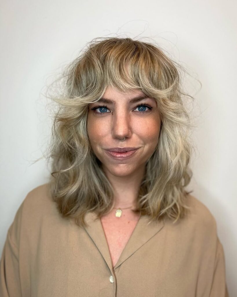 A woman with a blonde wavy shag haircut with curtain bangs and soft layers.