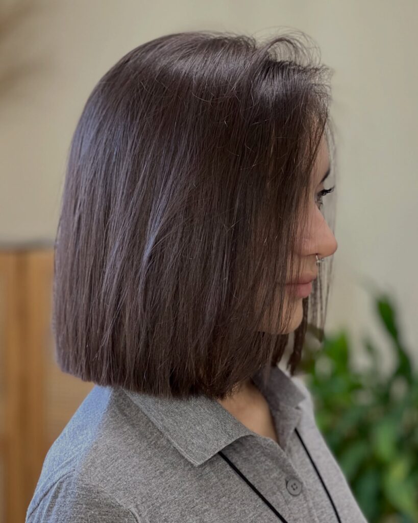 A woman with a straight neck length mocha brunette bob featuring a blunt cut and a smooth glossy finish is seen from a side profile.