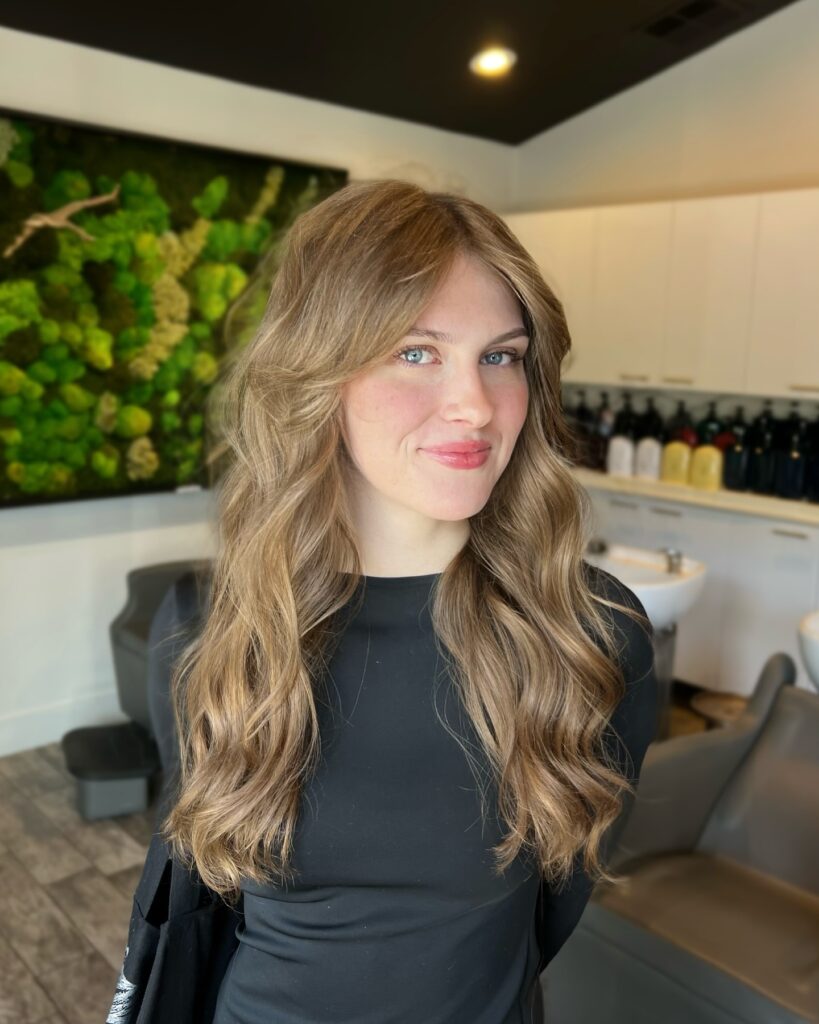 A young woman with long wavy honey blonde hair and face framing layers smiling in a salon setting.