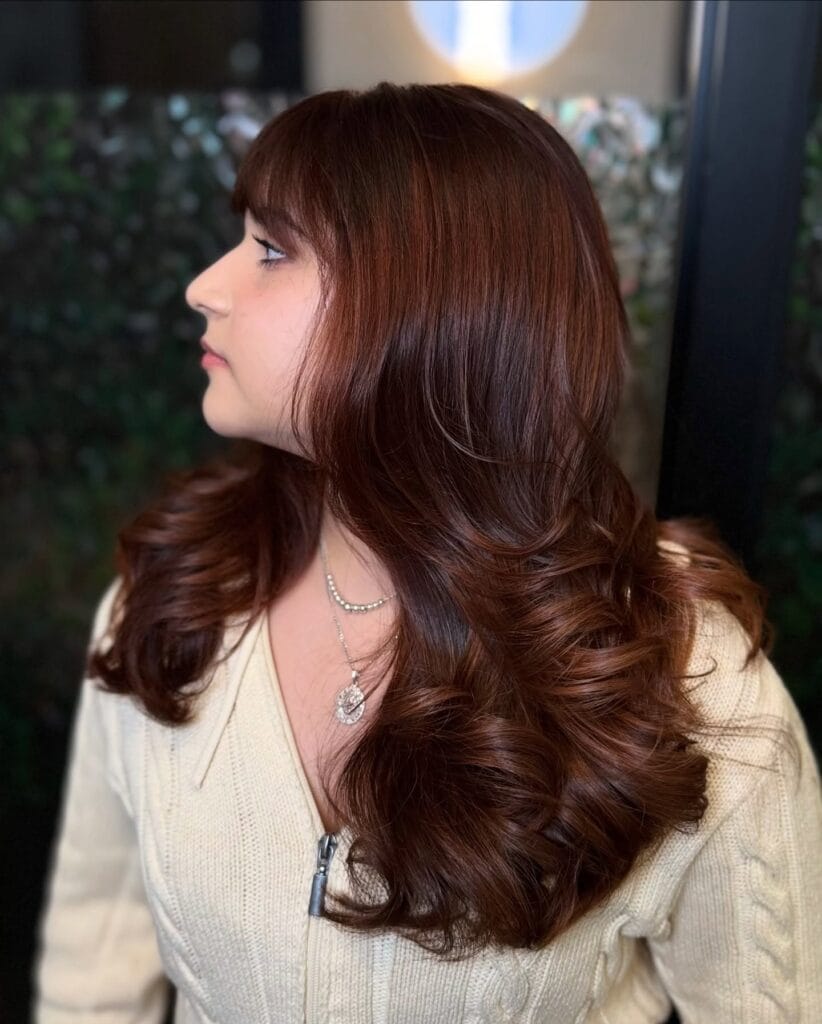 A woman with long glossy auburn hair featuring full straight bangs and bouncy curls at the ends.